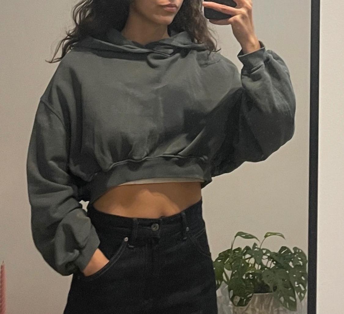 Woman taking a mirror selfie wearing a grey cropped sweatshirt with puff sleeves and black jeans. White background with a green plant.