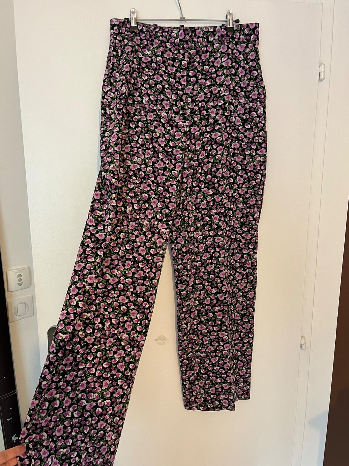 Wide-leg trousers with a purple floral print on a black background, hanging on a hanger on a white door. Casual and relaxed style.
