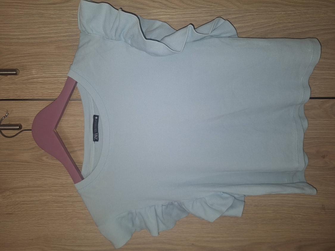 Ribbed light blue T-shirt with frill sleeves on a wooden surface. A pink hanger and the label with the text ZARA are visible.