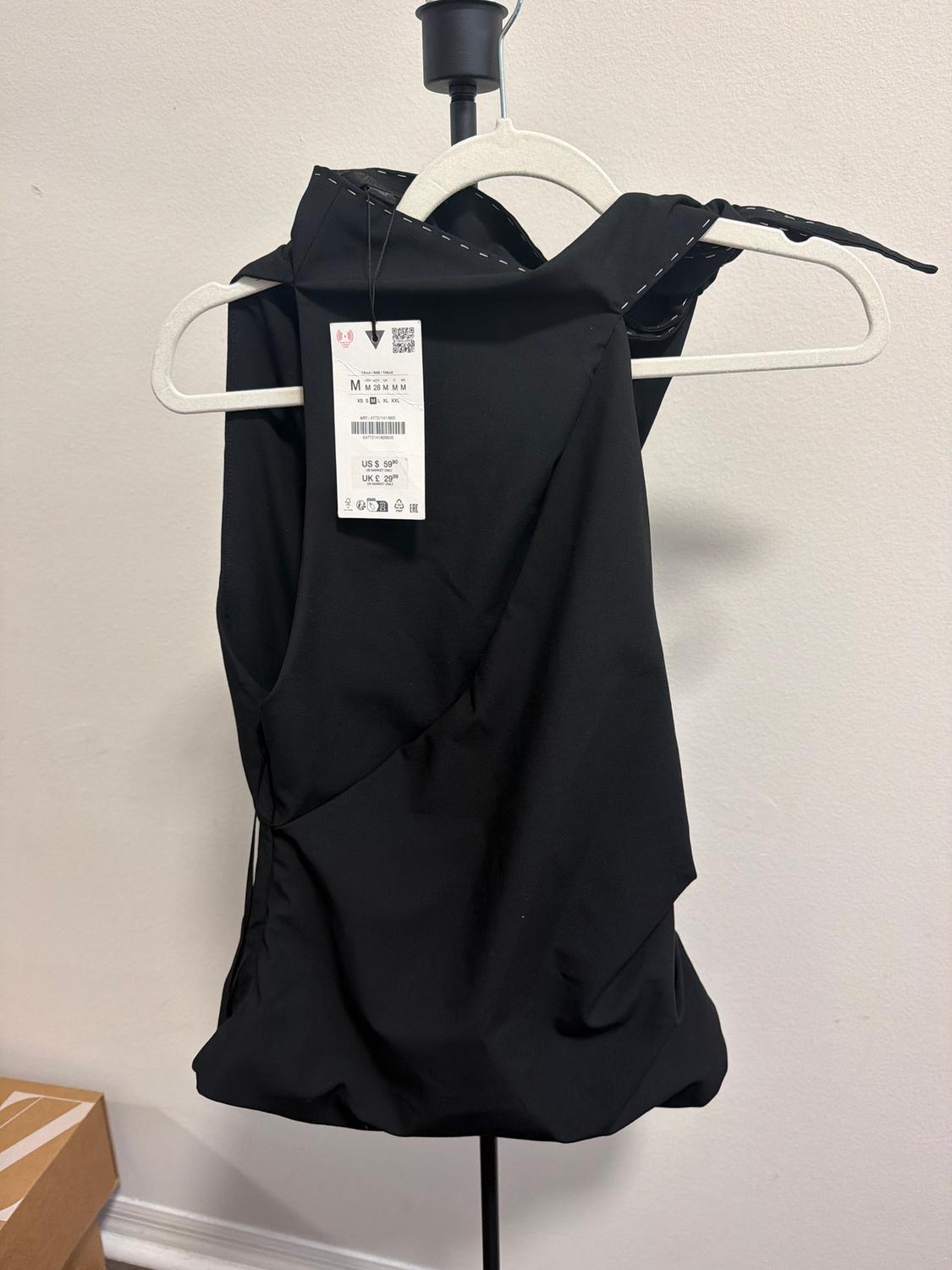 Black sleeveless top on a white hanger with a visible price tag. White background and brown box in the bottom left corner.