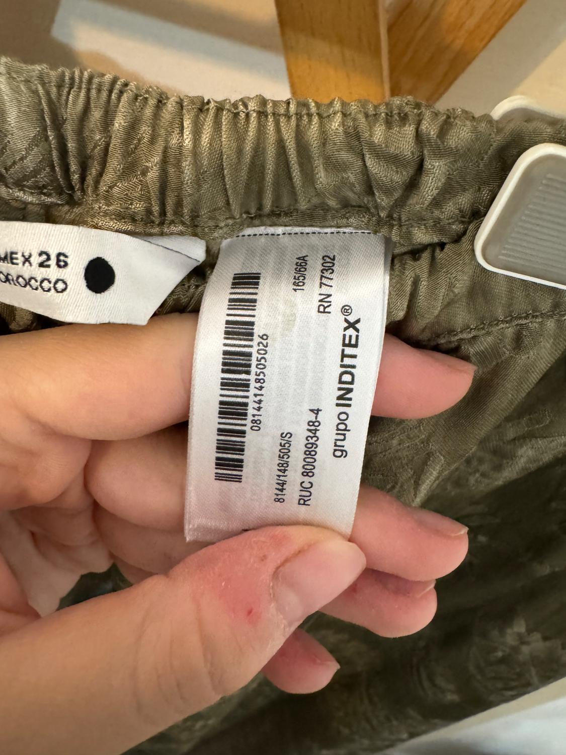 Χέρι κρατά πράσινες ετικέτες ρούχων. Κείμενο: MEX 26, 08144148505026, 8144/148/505/S, RUC 80089348-4, 165/66A, RN 77302, όμιλος INDITEX®