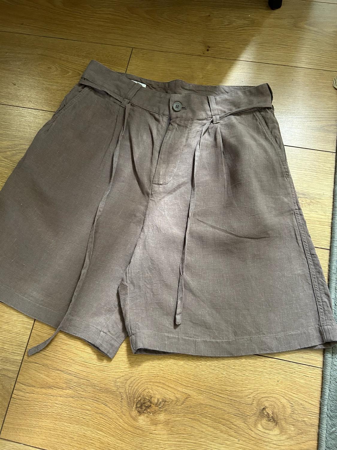 Brown tailored shorts with pleats and matching thin belt, laid out on a wooden floor illuminated by natural light. Relaxed style.
