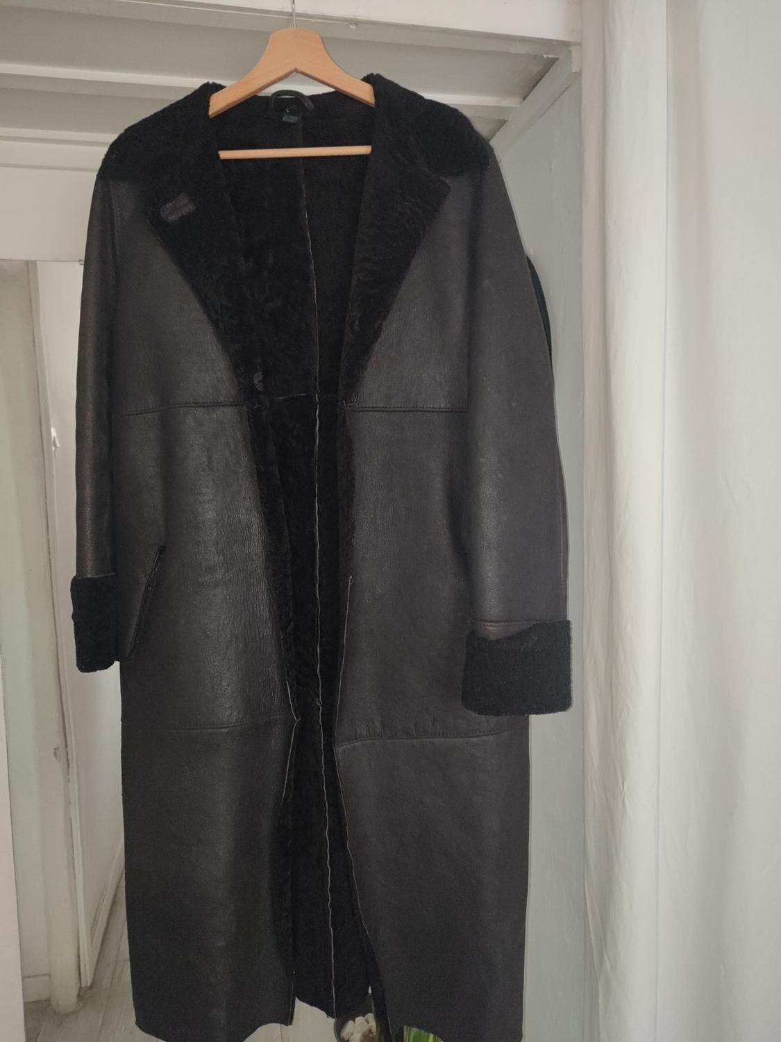Black long coat hanging on a wooden hanger, with a soft-textured interior and cuffs, in a well-lit indoor setting.