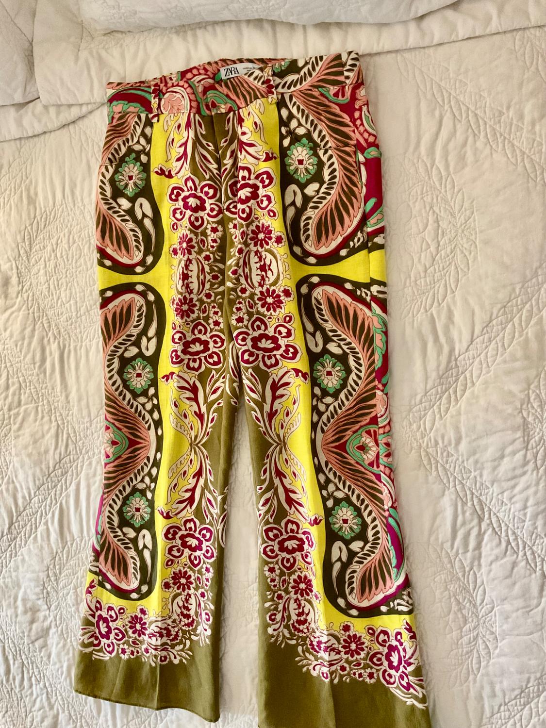 Pants with an eye-catching floral print in yellow, olive green, and maroon, on a textured white background. ZARA label at the waist.