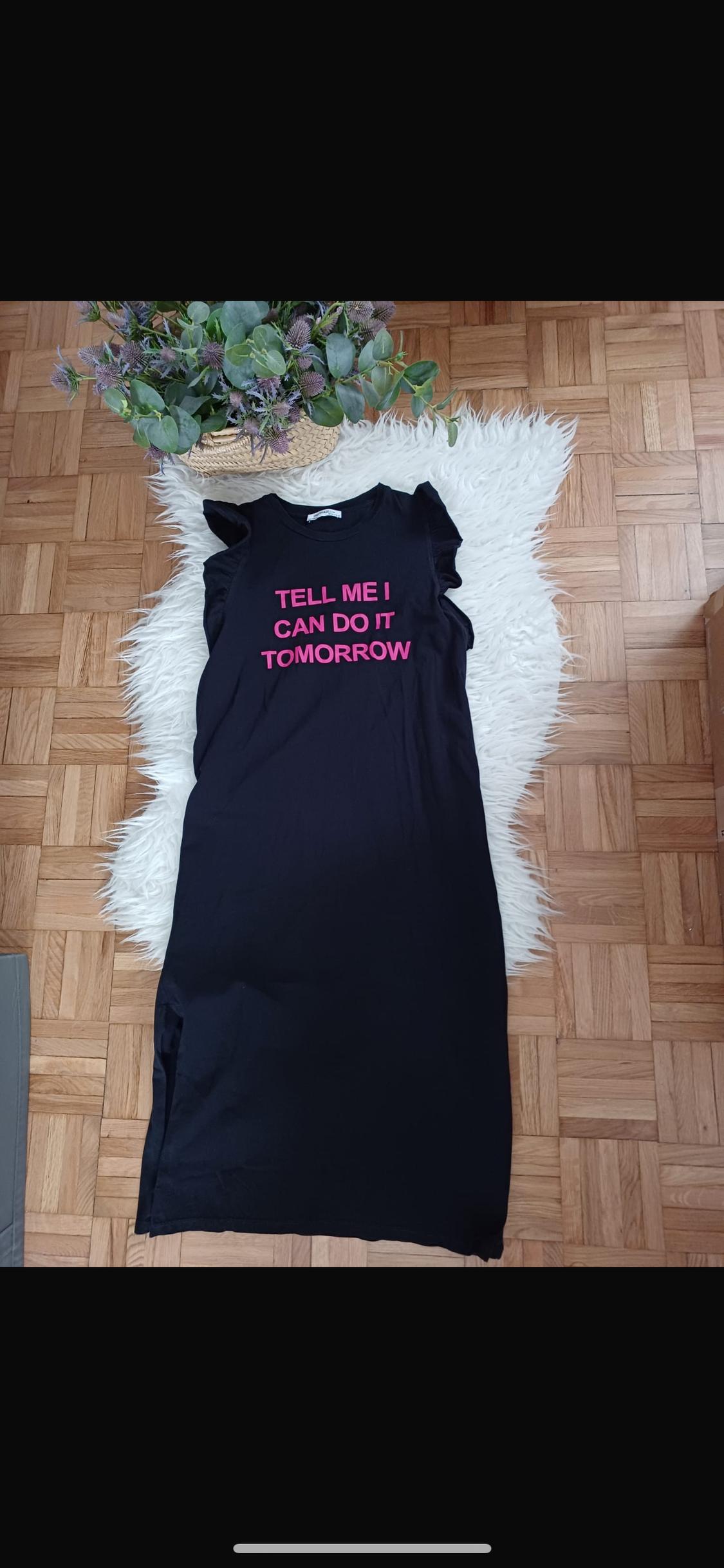 Μαύρο φόρεμα με ροζ κείμενο "Tell me I can do it tomorrow" πάνω σε λευκό χαλί, δίπλα σε φυτό σε γλάστρα πάνω σε ξύλινο δάπεδο.