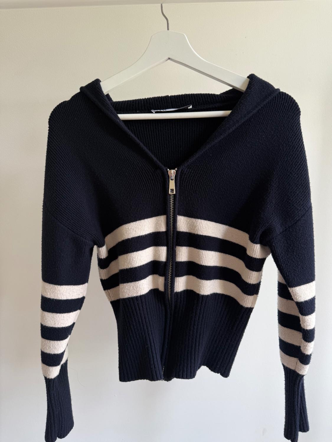 Navy knitted jacket with front zip fastening, hood and horizontal white stripes on torso and sleeves, hanging on a white hanger.