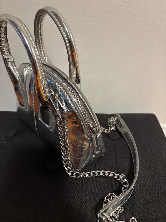 Small silver handbag with crocodile texture, rigid handles, and metallic chain on a black background.