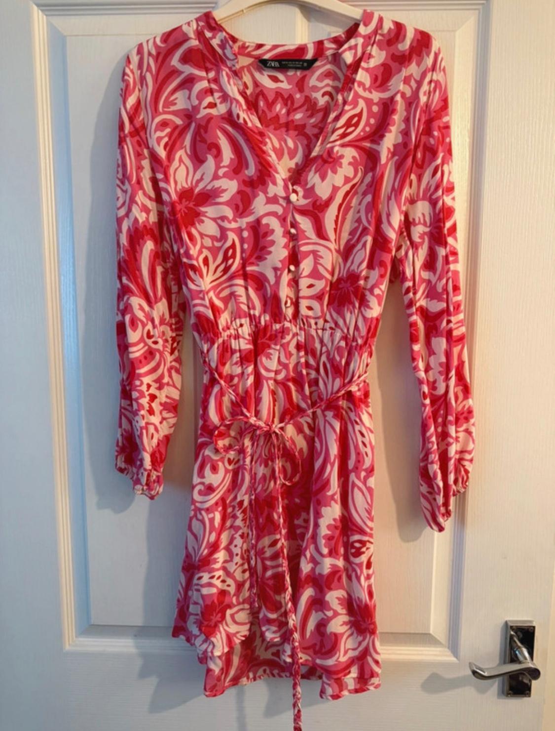 Dress hanging on a door, with a floral print in pink and white tones, long sleeves, and an adjustable belt.
