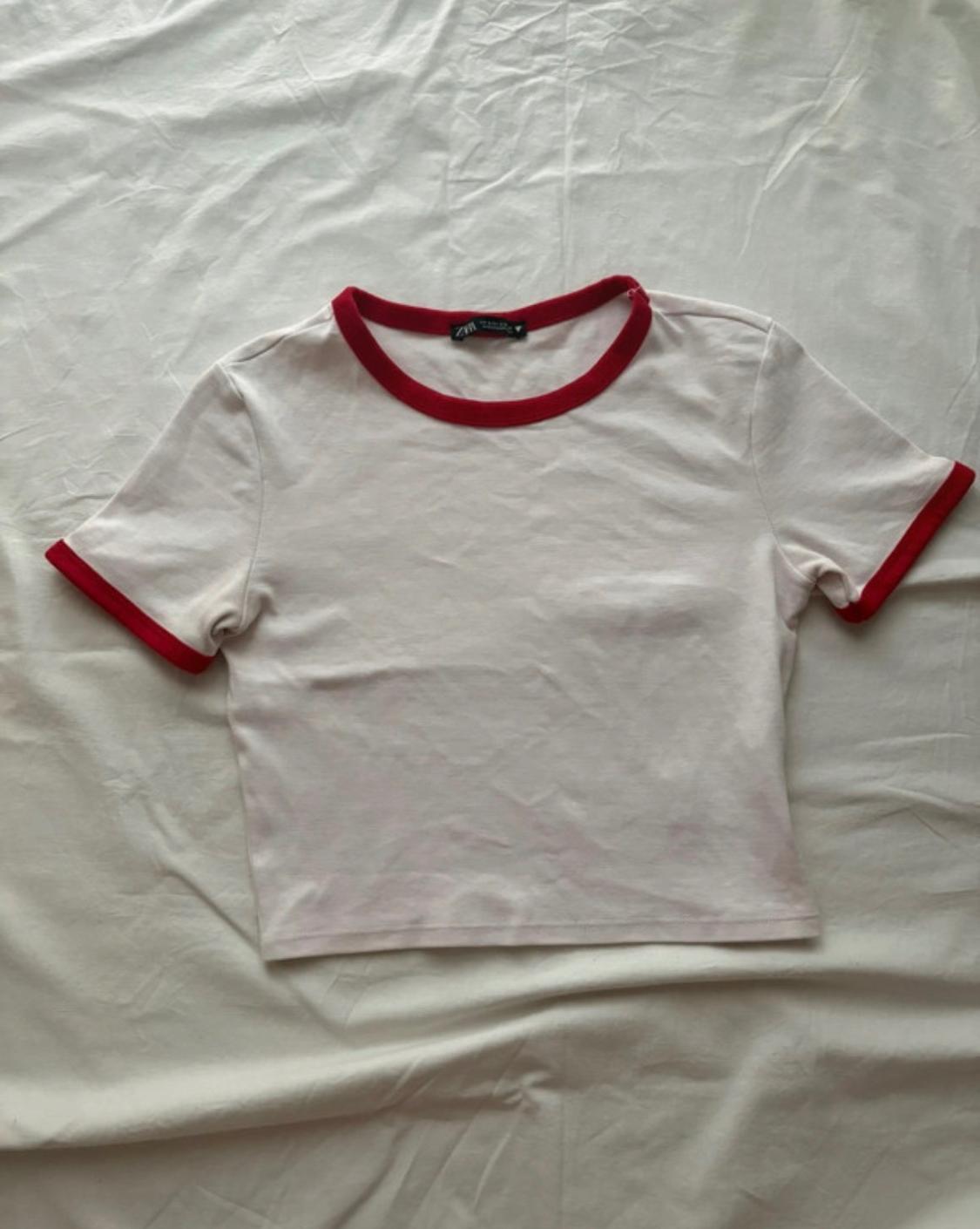 White short-sleeved T-shirt with red trim around the collar and sleeves, on a crumpled white sheet.