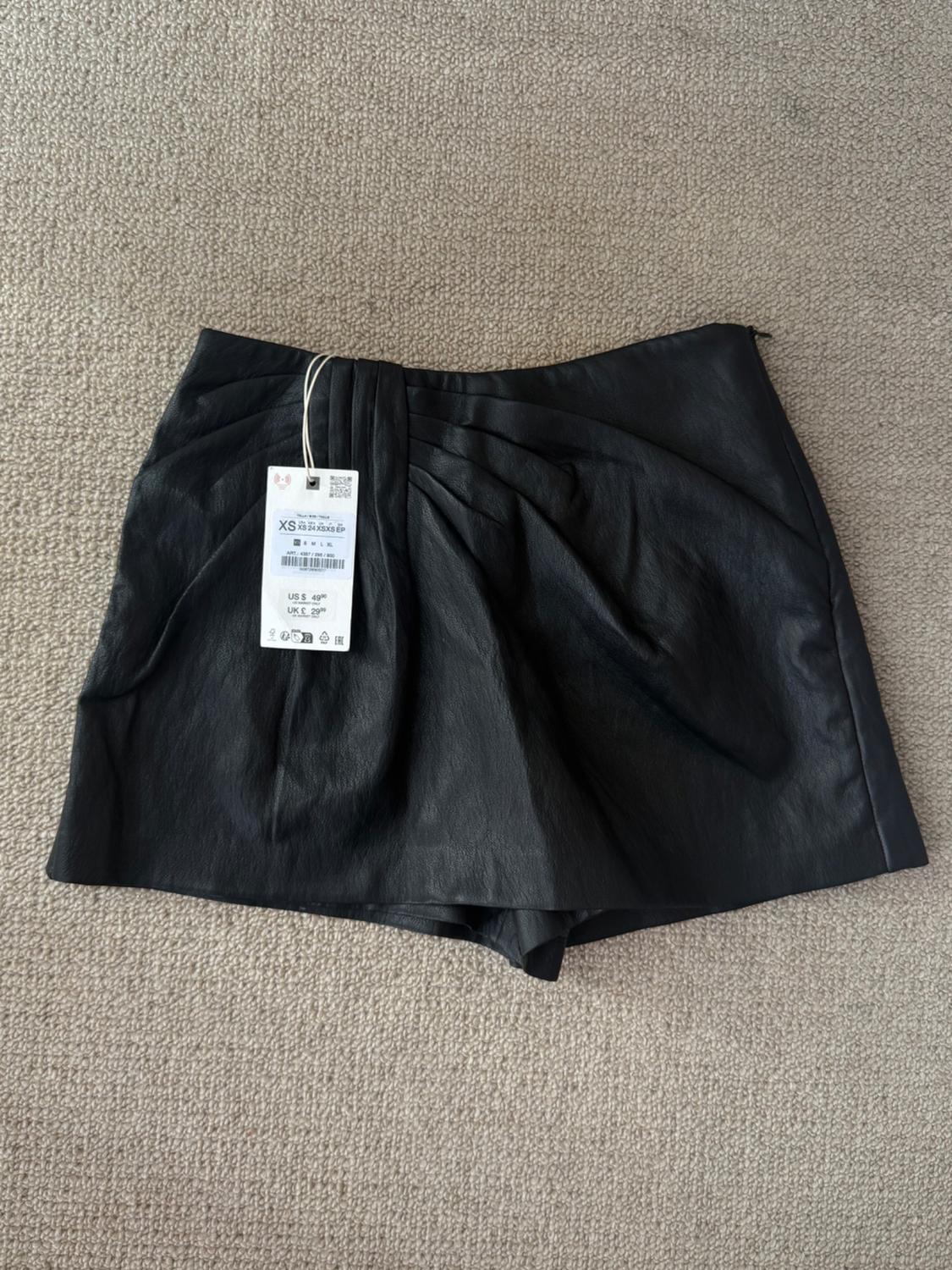 Black shorts on a beige carpet. Label with text: XS, ART: 4387 / 255 / 800, US $ 49.90, UK £ 29.99.
