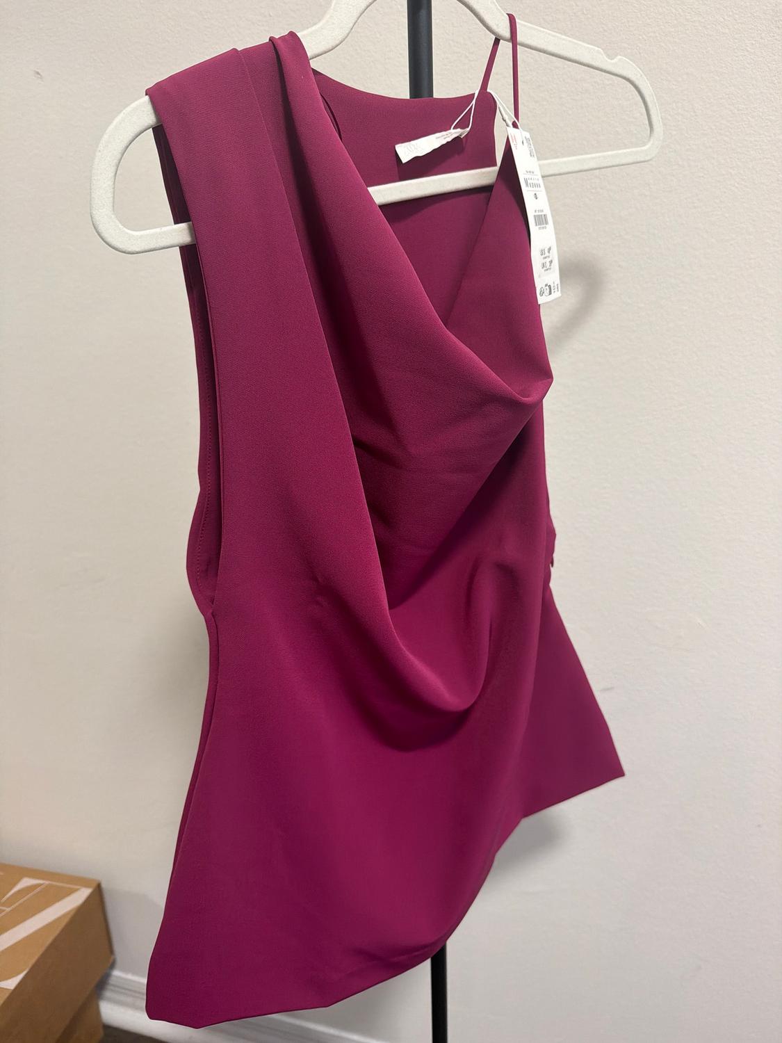 Fuchsia sleeveless blouse with a draped neckline, hanging on a white hanger. Visible tag on the right strap. White background.