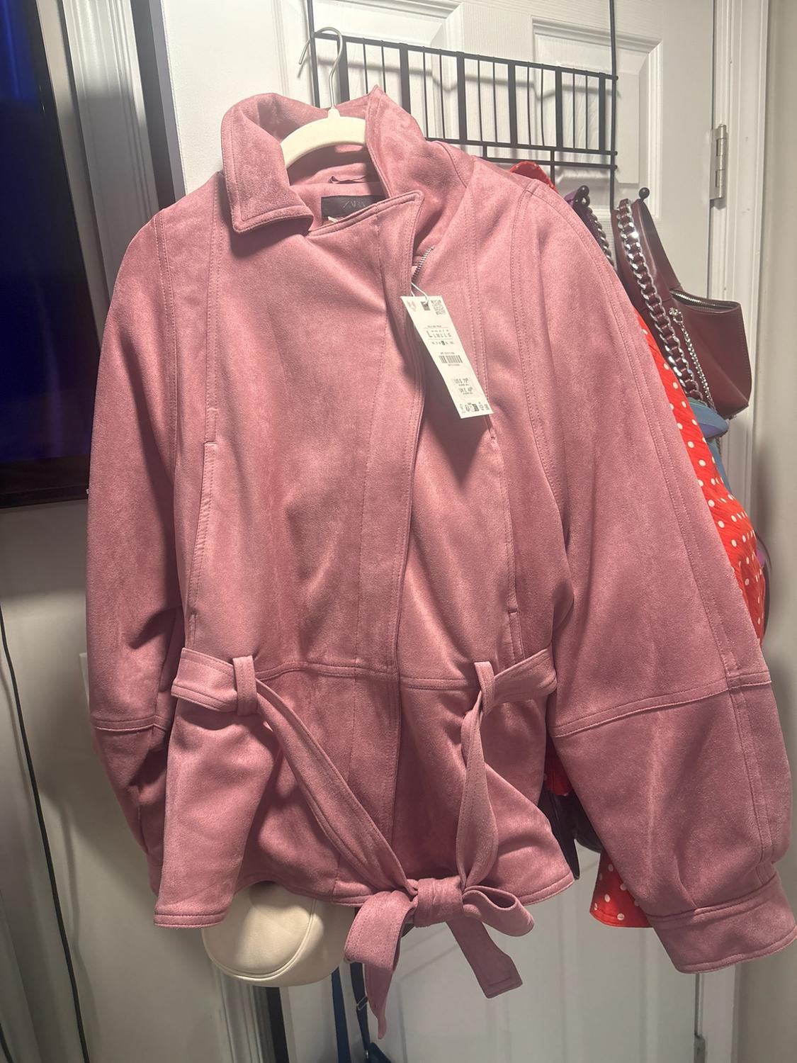 Pink faux suede jacket with a collar and tied belt, hanging on a hanger on a white door.