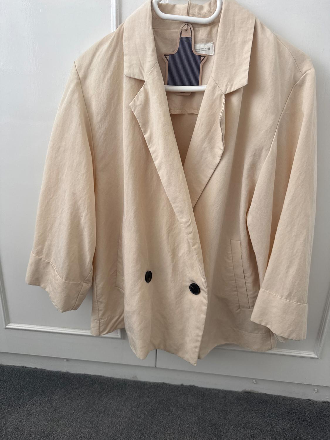 Beige long-sleeved jacket hanging on a white hanger, with black buttons, over a white door.