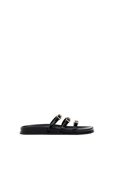 Black flat sandal with three straps and gold buckles, profile view at the bottom of the image against a white background.
