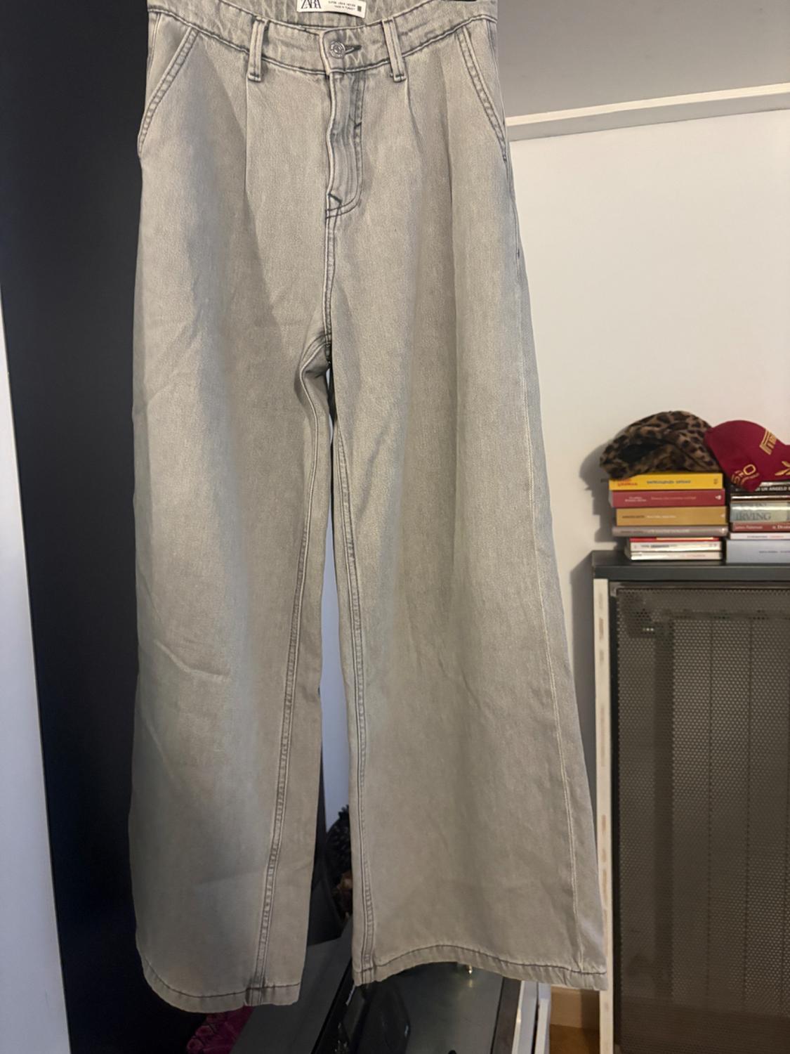 Wide grey denim trousers hanging on a clothes rail, with a background of stacked books and a red cap.