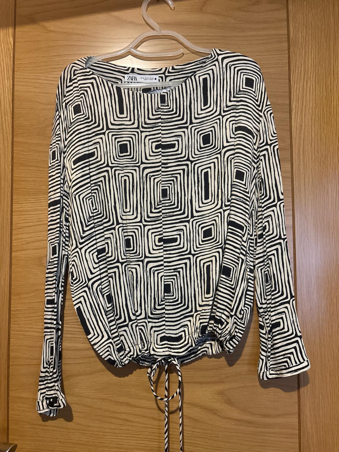 Long-sleeved blouse with a black and white geometric print, adjustable drawstring hem, hanging on a wooden door. Text: ZARA.