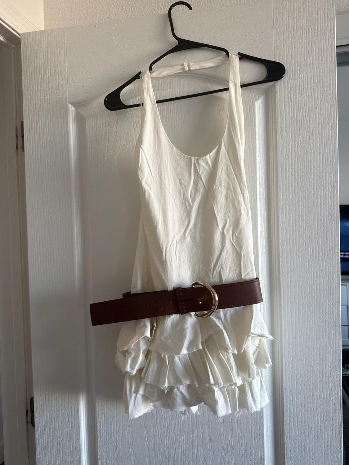 Short white ruffled dress hanging on a white door, paired with a wide brown belt with a gold buckle.