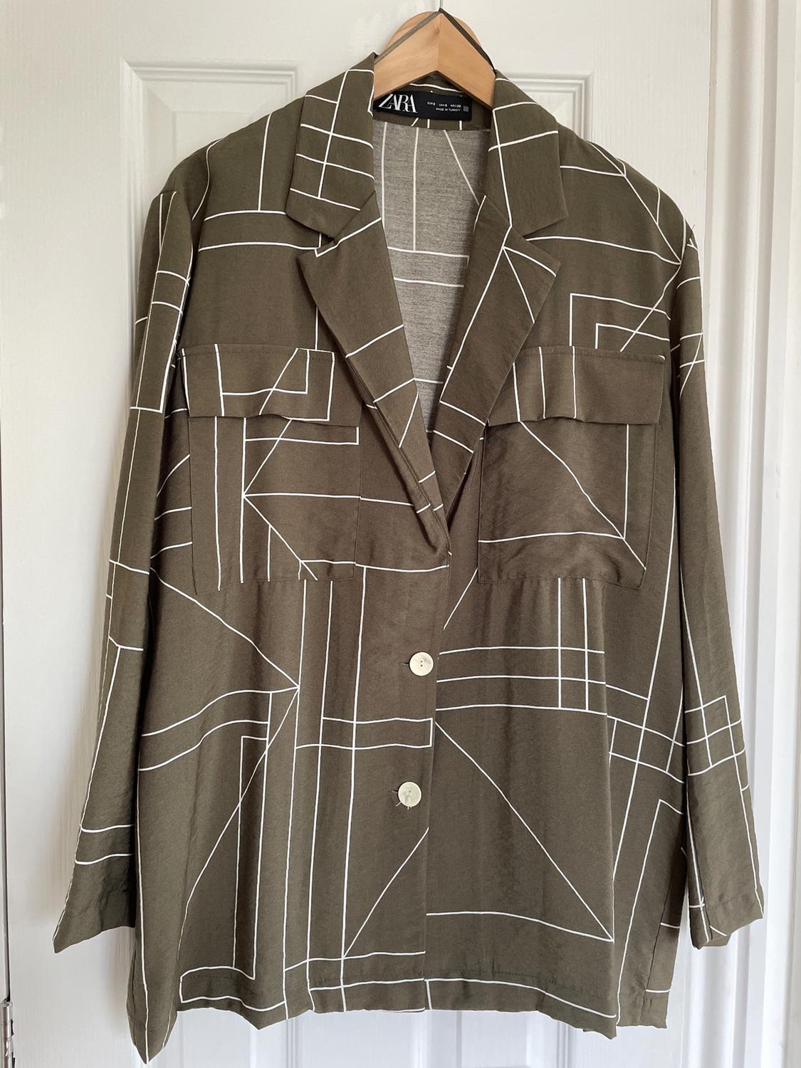 Olive green jacket with a white geometric pattern, front pockets, and light-coloured buttons, hanging on a wooden hanger.