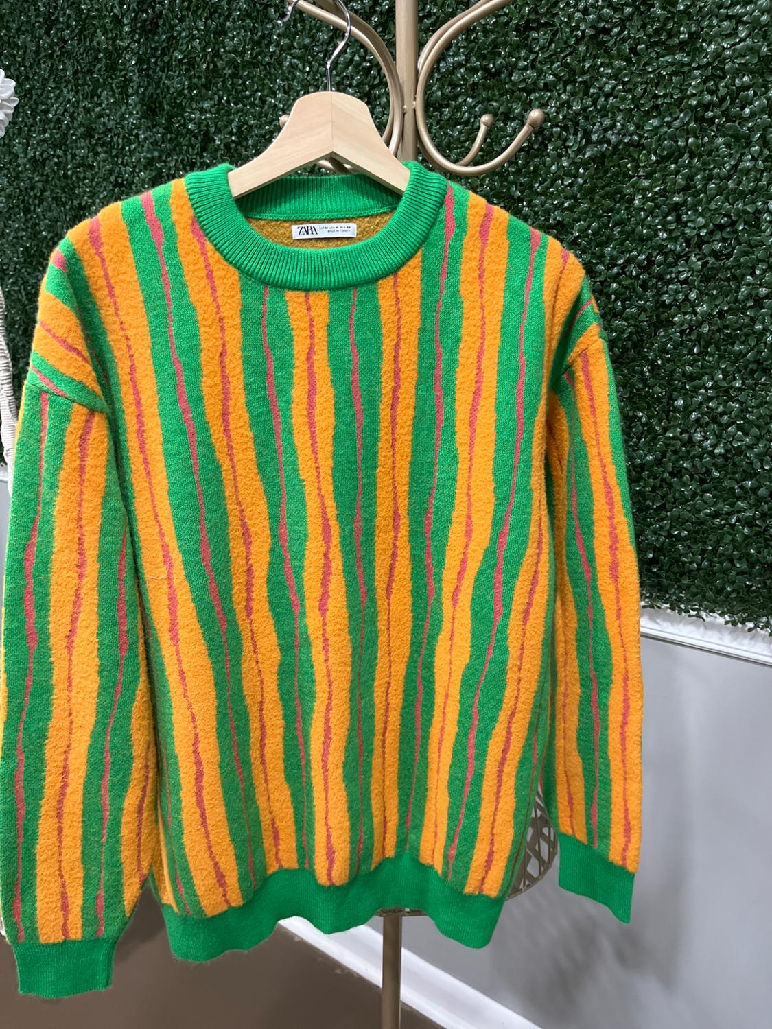 Sweater hanging with green, yellow, and pink vertical stripes. Plant wall background. Visible tag on the collar.