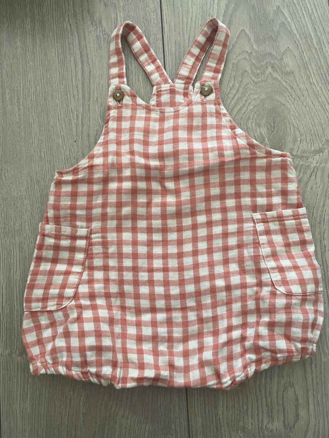 Children's dungarees in white and pink check with straps and front pockets, on a light wooden floor.