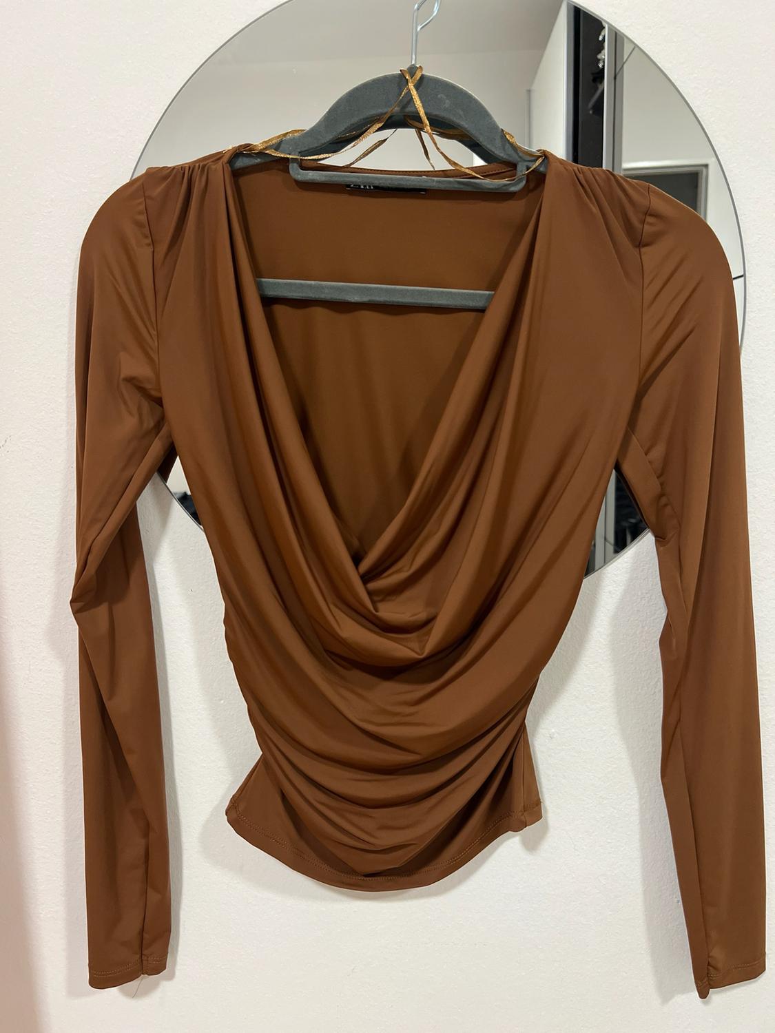 Brown long-sleeved top with a draped, gathered neckline, hanging on a hanger in front of a round mirror on a white wall.