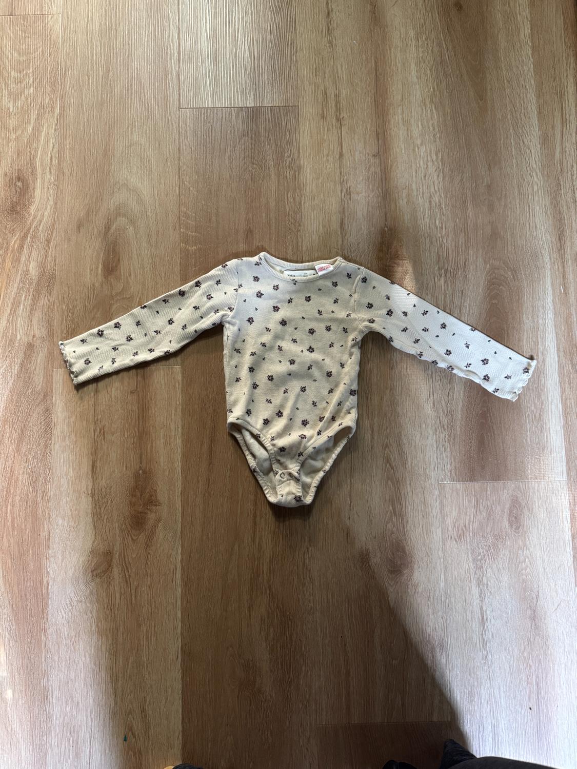 White long-sleeved bodysuit with a small black floral print, on a light wooden floor.