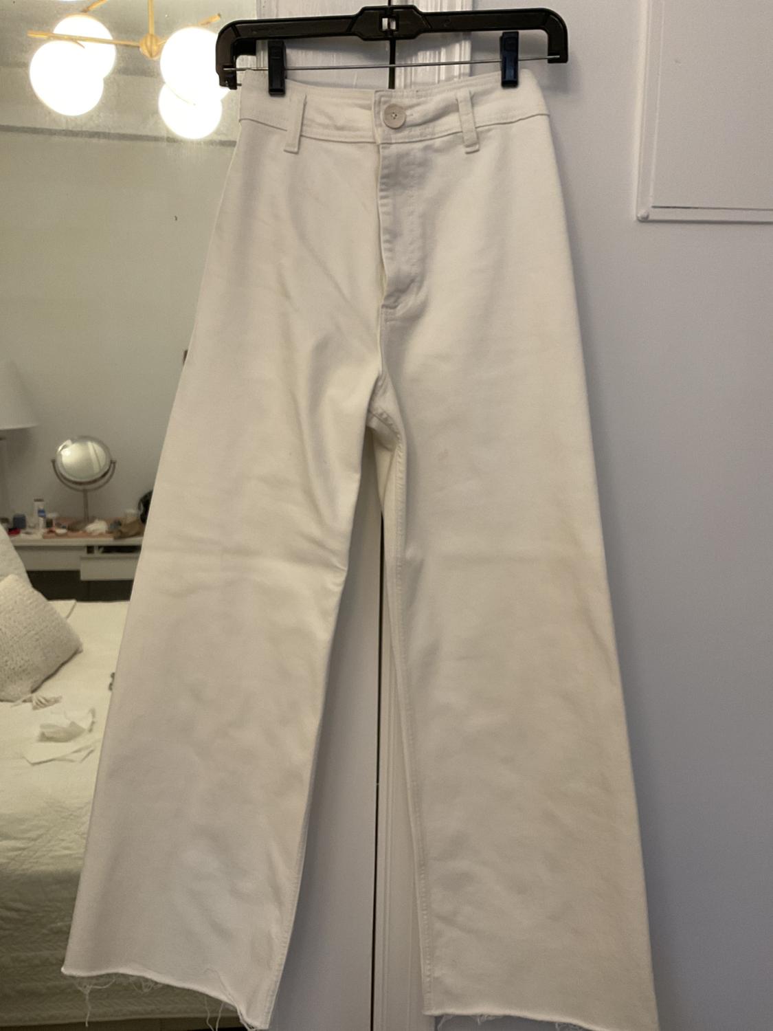 High-waisted white pants, wide-leg with a frayed hem, hanging on a black hanger in a bedroom with a bed and mirror in the background.