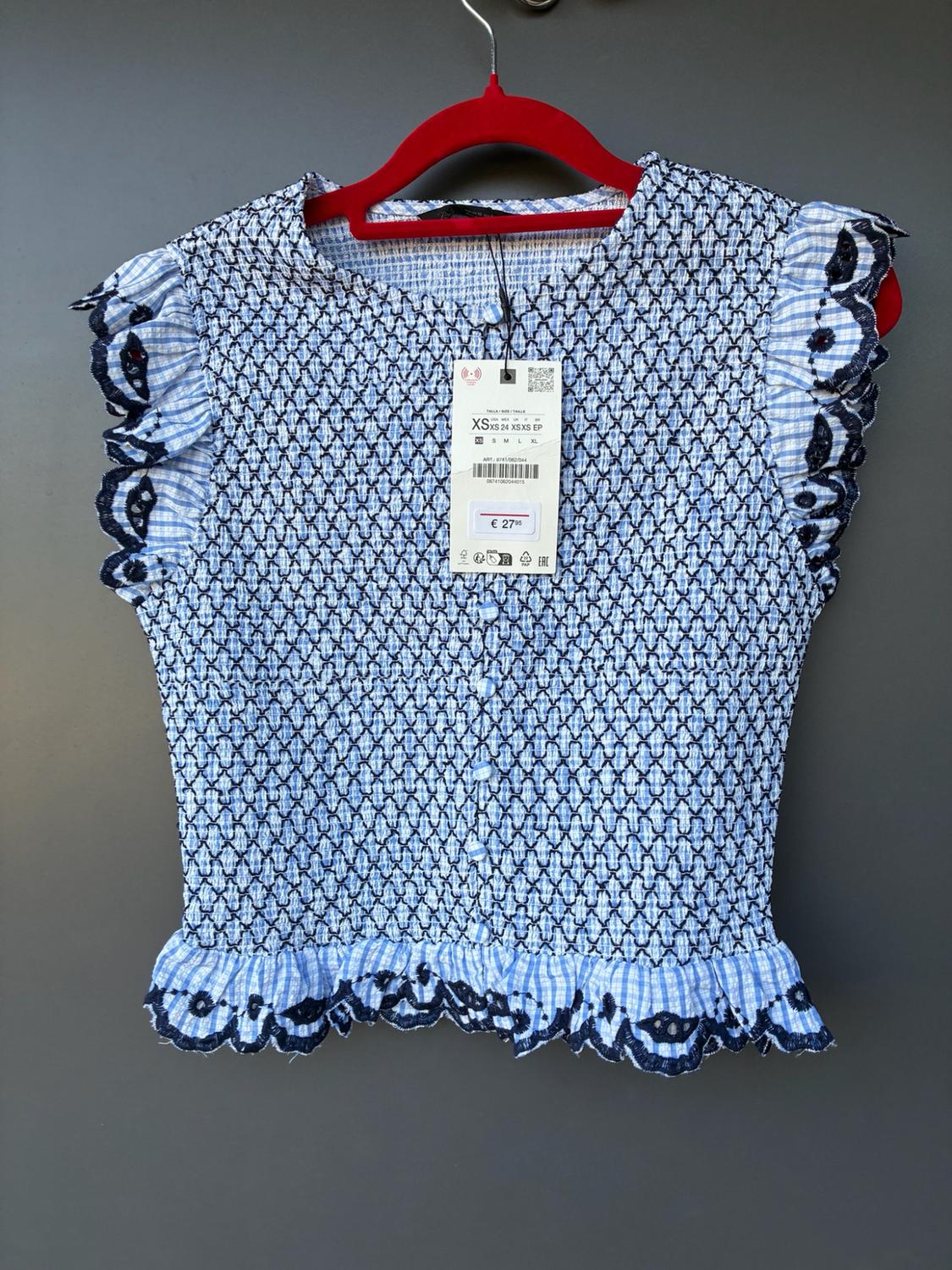 Blue and white checked shirred top with embroidered frills on a red hanger. Text: XS XS 24 XSXS EP, S M L XL, €27.95.