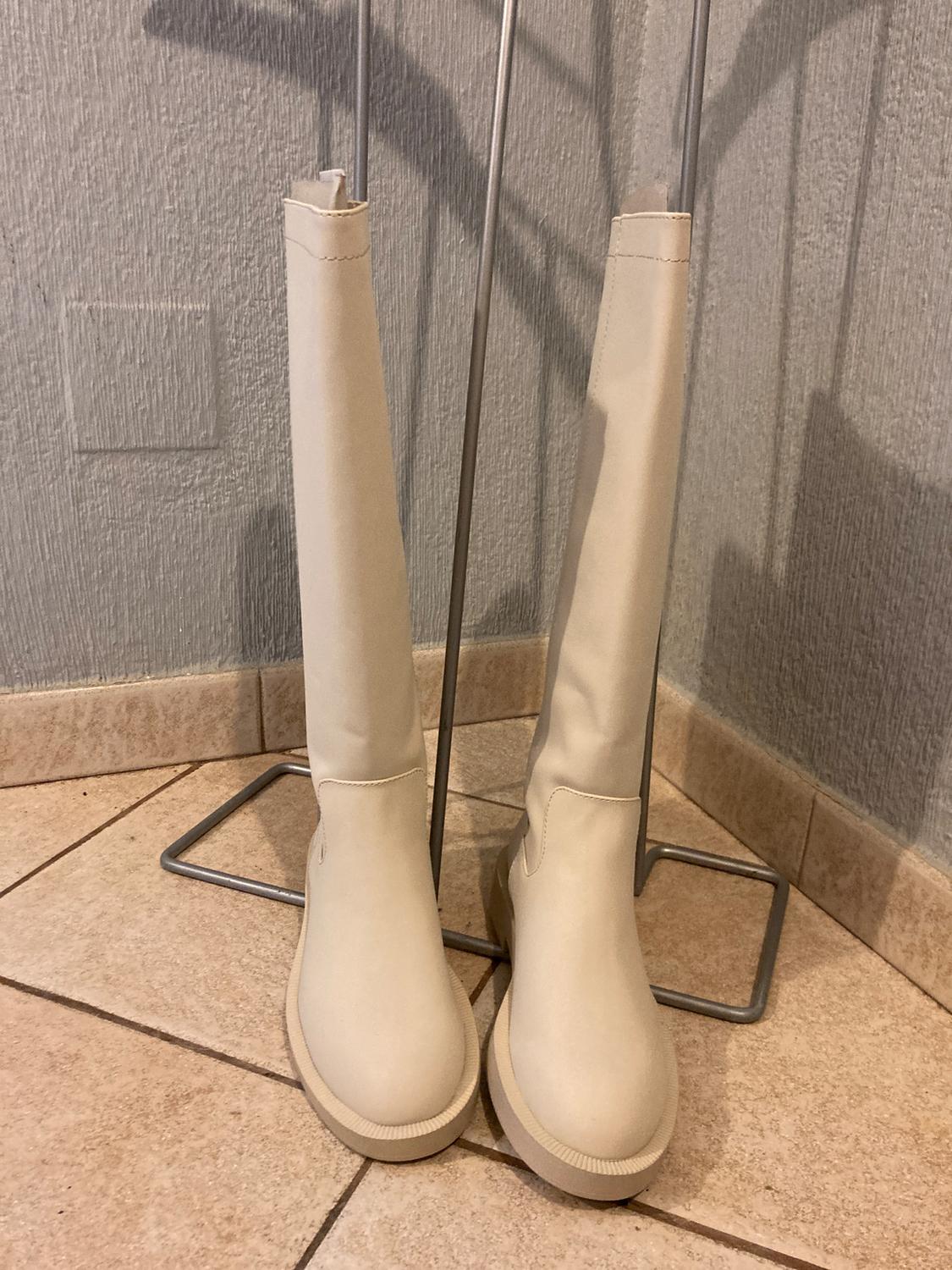 Beige knee-high boots on a metal stand on a beige tiled floor. Grey textured wall in the background.