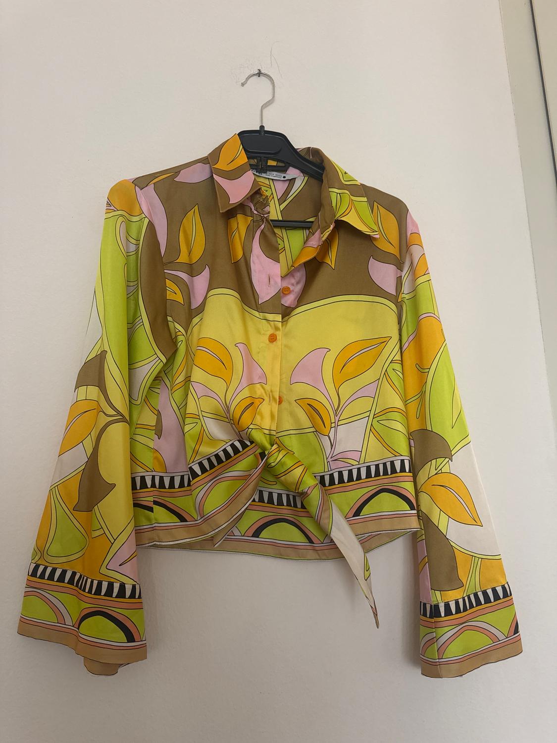 Colourful blouse with a floral print in shades of green, yellow, and pink, hanging on a hanger against a white wall.