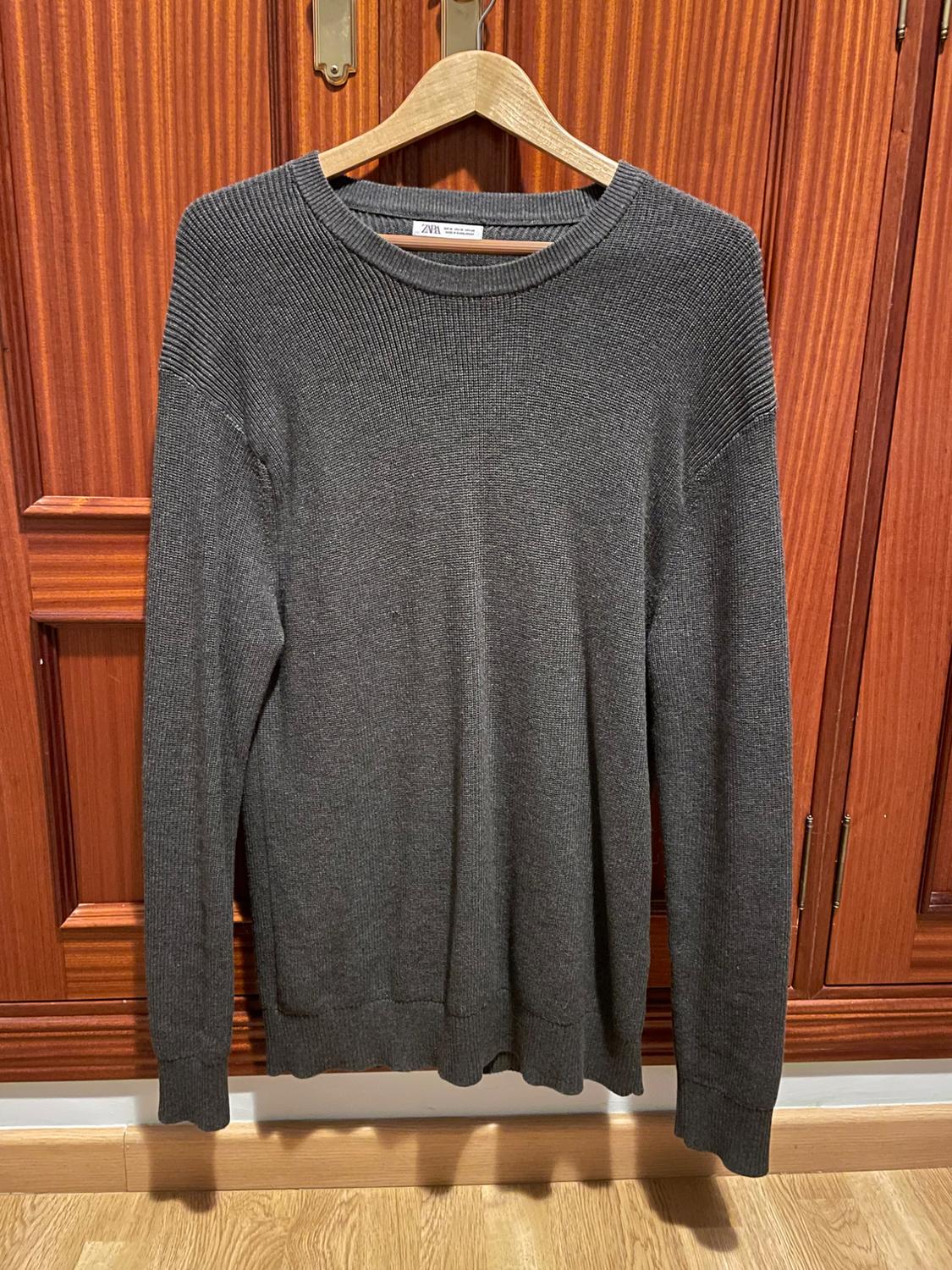 Grey knitted jumper hanging on a wooden hanger in front of a wooden wardrobe. Visible label: "ZARA".