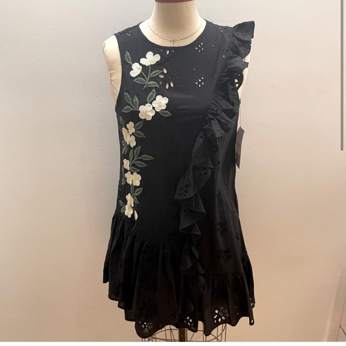 Front view of a black dress with white floral embroidery and ruffles on the side.