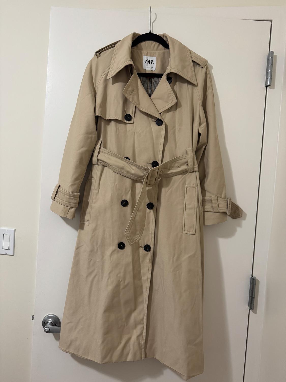 Beige trench coat with a belt and black buttons, hanging on a white door. Visible tag with the text "Zara".