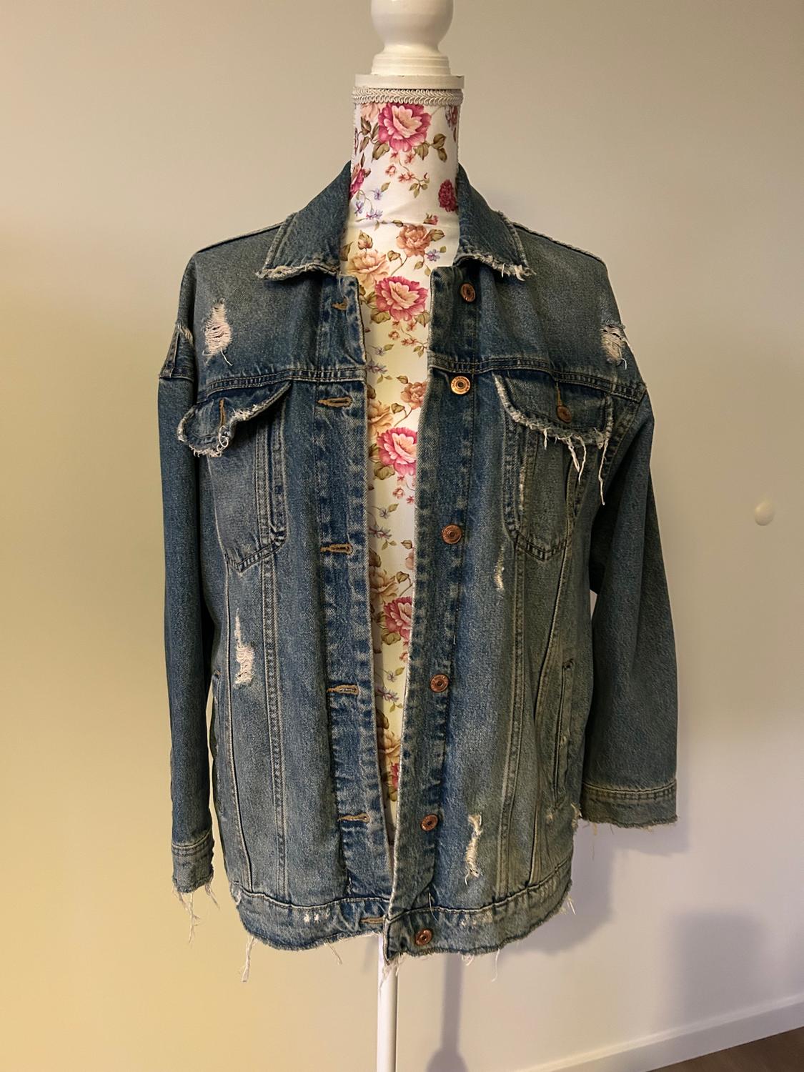 Distressed denim jacket on a mannequin with a floral print. Neutral background and visible brown buttons.