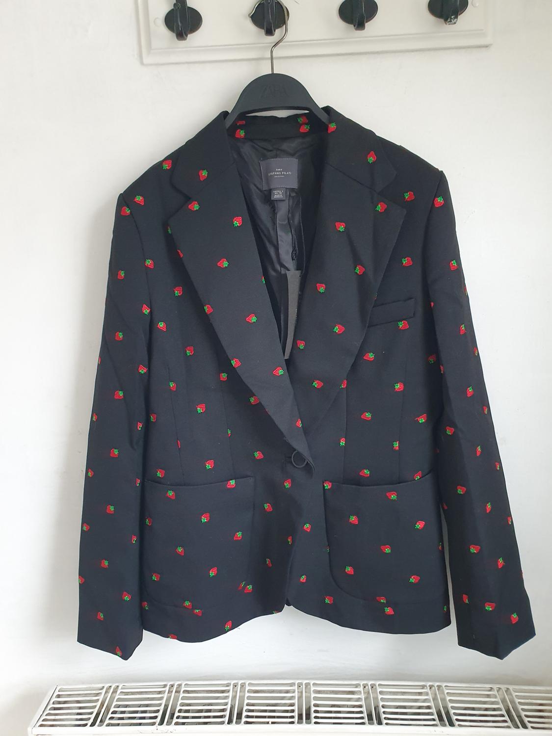 Black jacket with a small red and green strawberry print, hanging on a black hanger against a white wall.