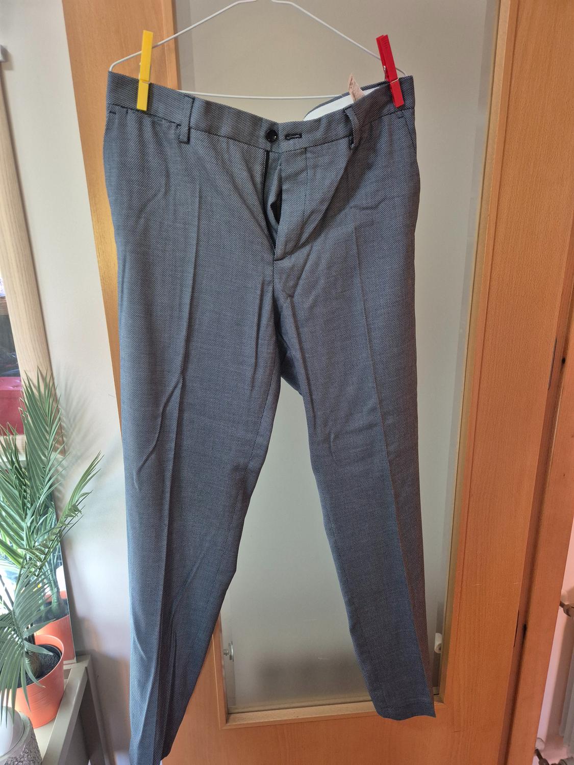 Grey trousers hanging on a hanger with colourful pegs, with a wooden door background and a potted plant on the left.