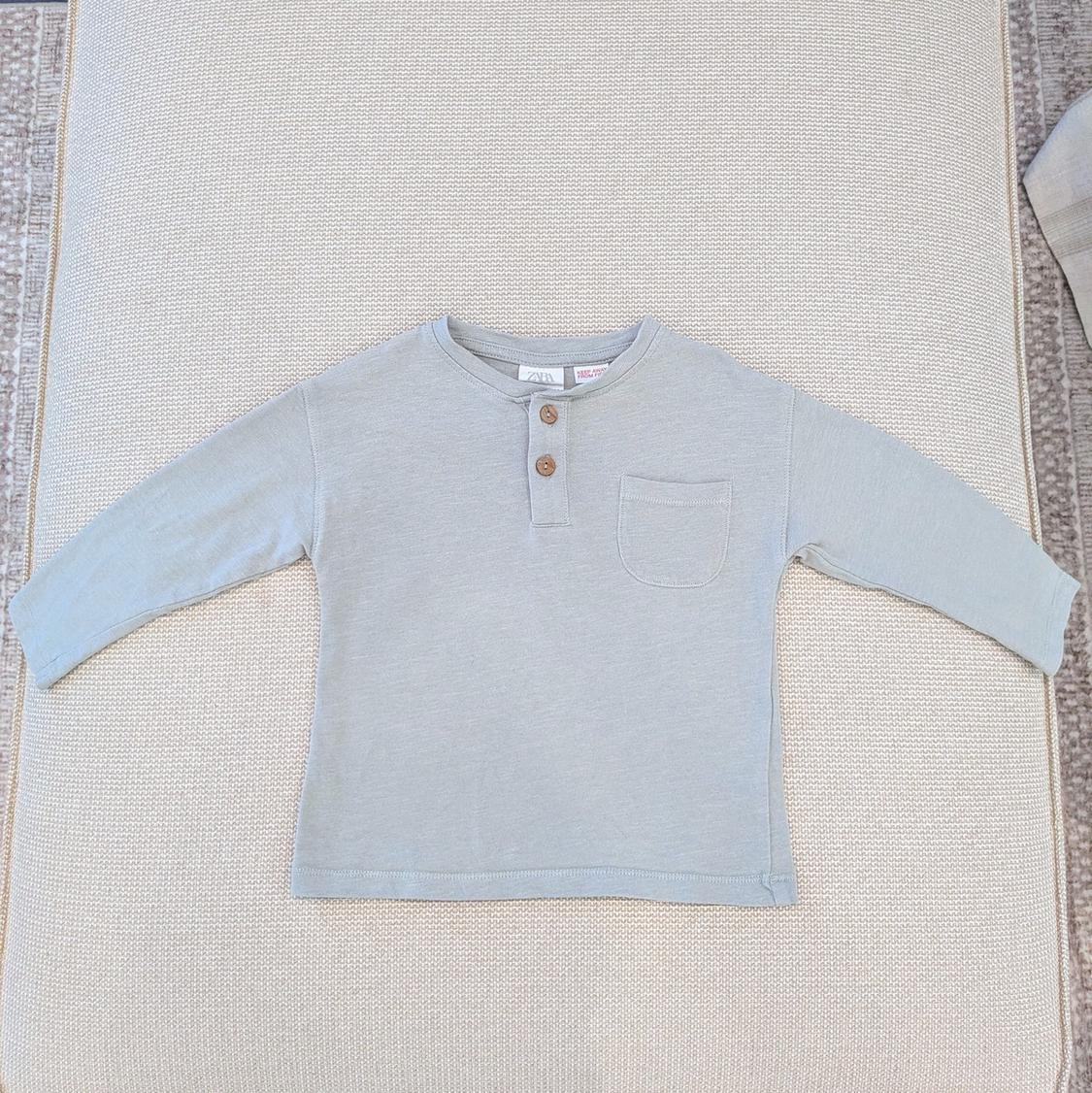 Light gray long-sleeve t-shirt with brown buttons and a chest pocket, on a textured beige surface.