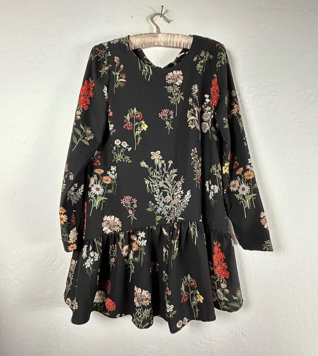 Black dress with colourful floral print, long sleeves and a flared skirt, hanging on a hanger against a white wall.