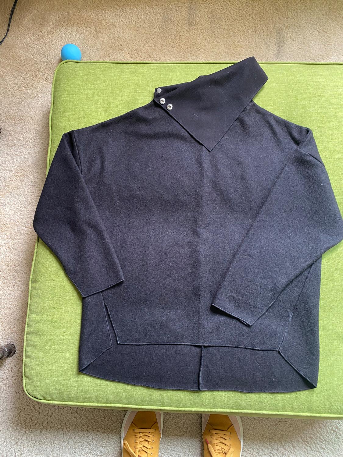 Black long-sleeve jersey with an asymmetrical collar and buttons, on a green cushion. Part of some yellow shoes visible below.
