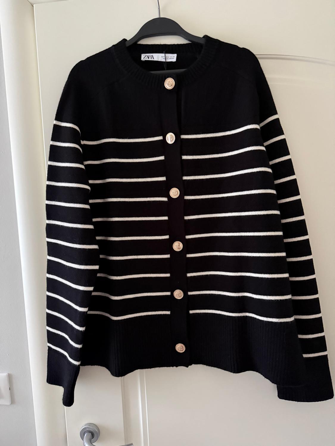 Black jacket with horizontal white stripes and gold buttons, hung on a black hanger over a white door.