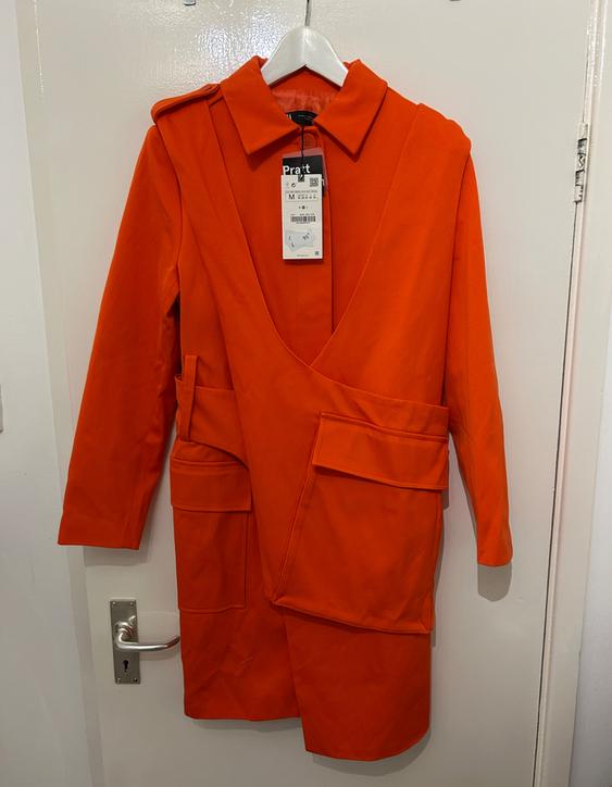 Orange coat with a double-breasted lapel and large pockets, hanging on a white door. Visible label with text "Pratt" and size M.