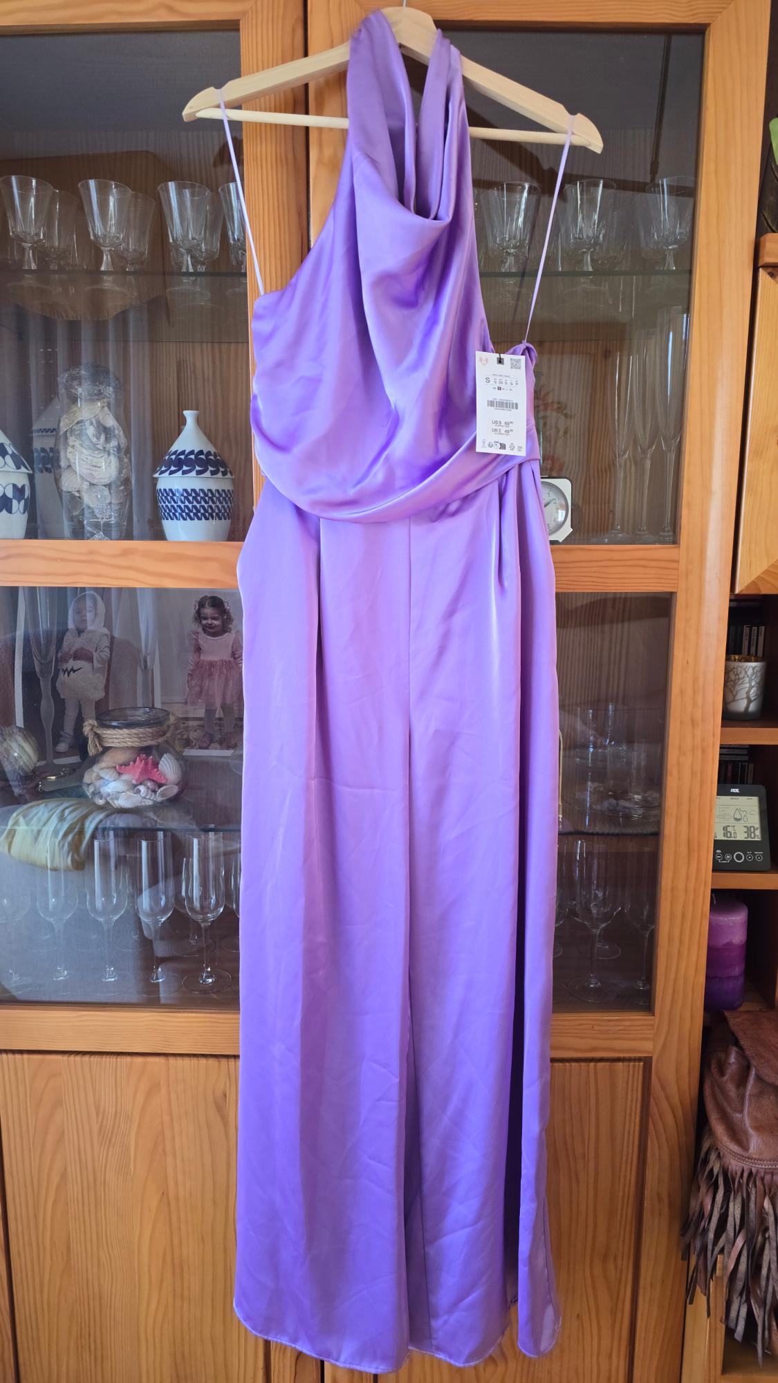 Lilac long dress hanging on a hanger in front of a wooden cabinet with crystal glasses and decorative vases.