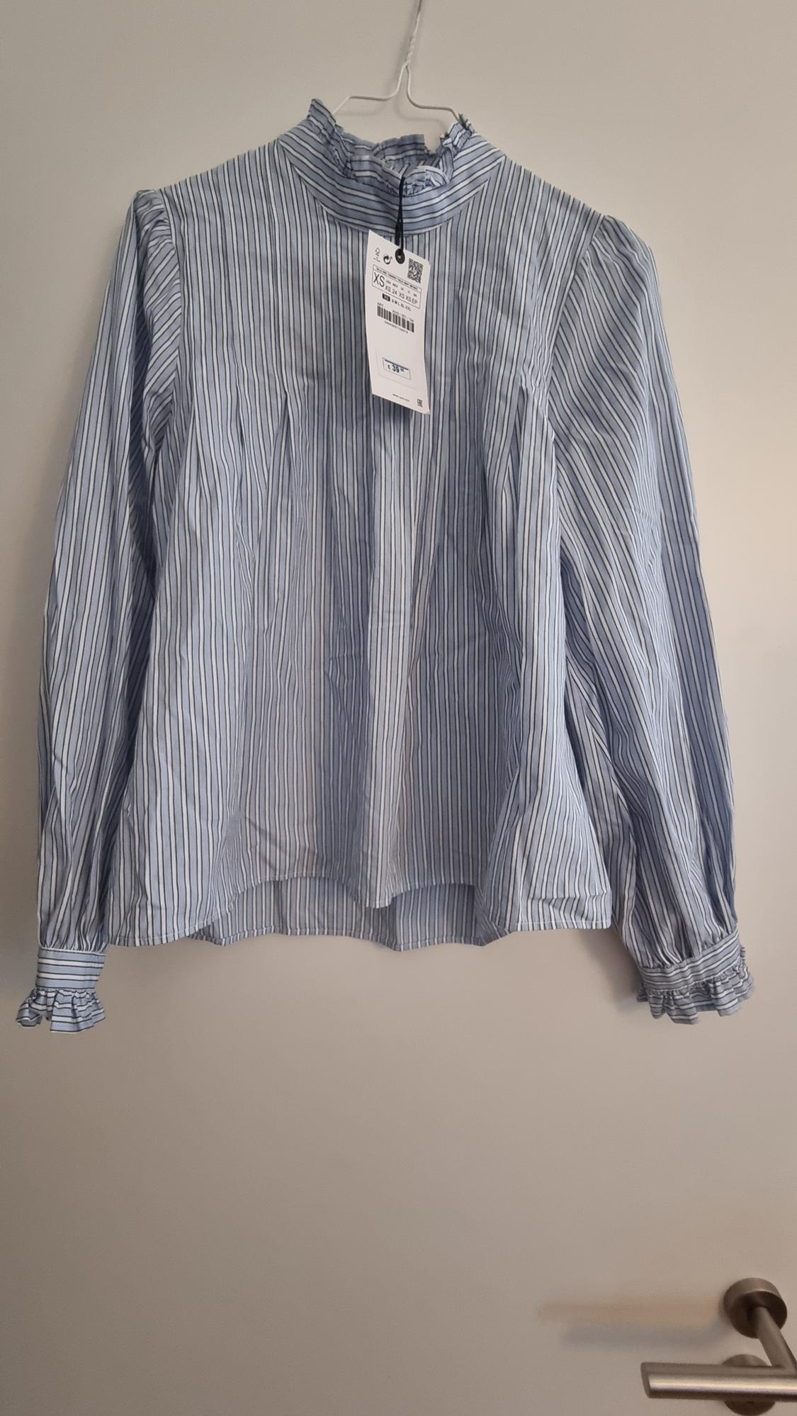 Blue and white striped blouse with a high neck and long sleeves, hanging on a hanger. Visible label with product details.