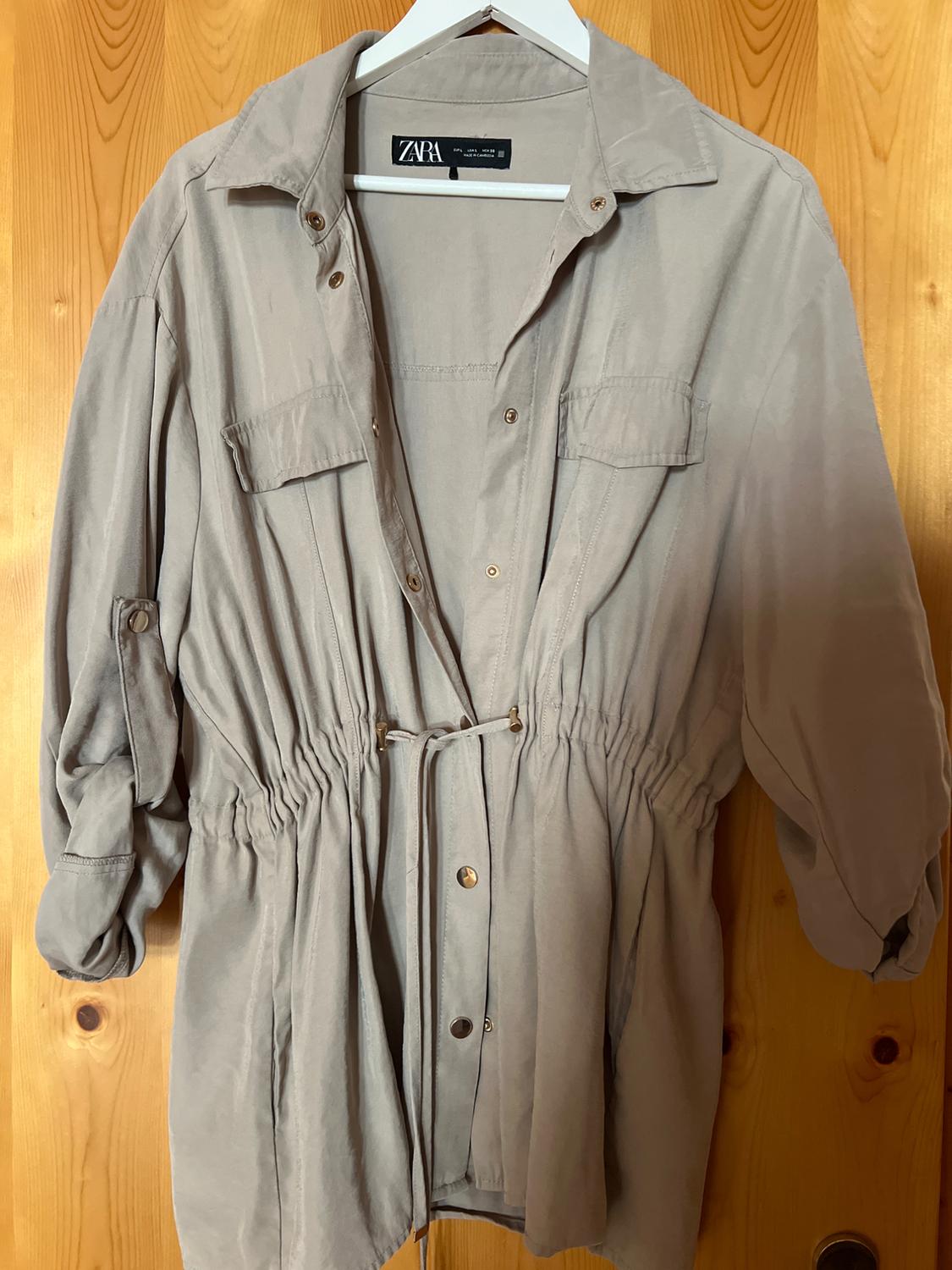 Beige shirt dress with ZARA label, hanging on a white hanger against a wooden background. Casual design with buttons, pockets and a fitted waist.