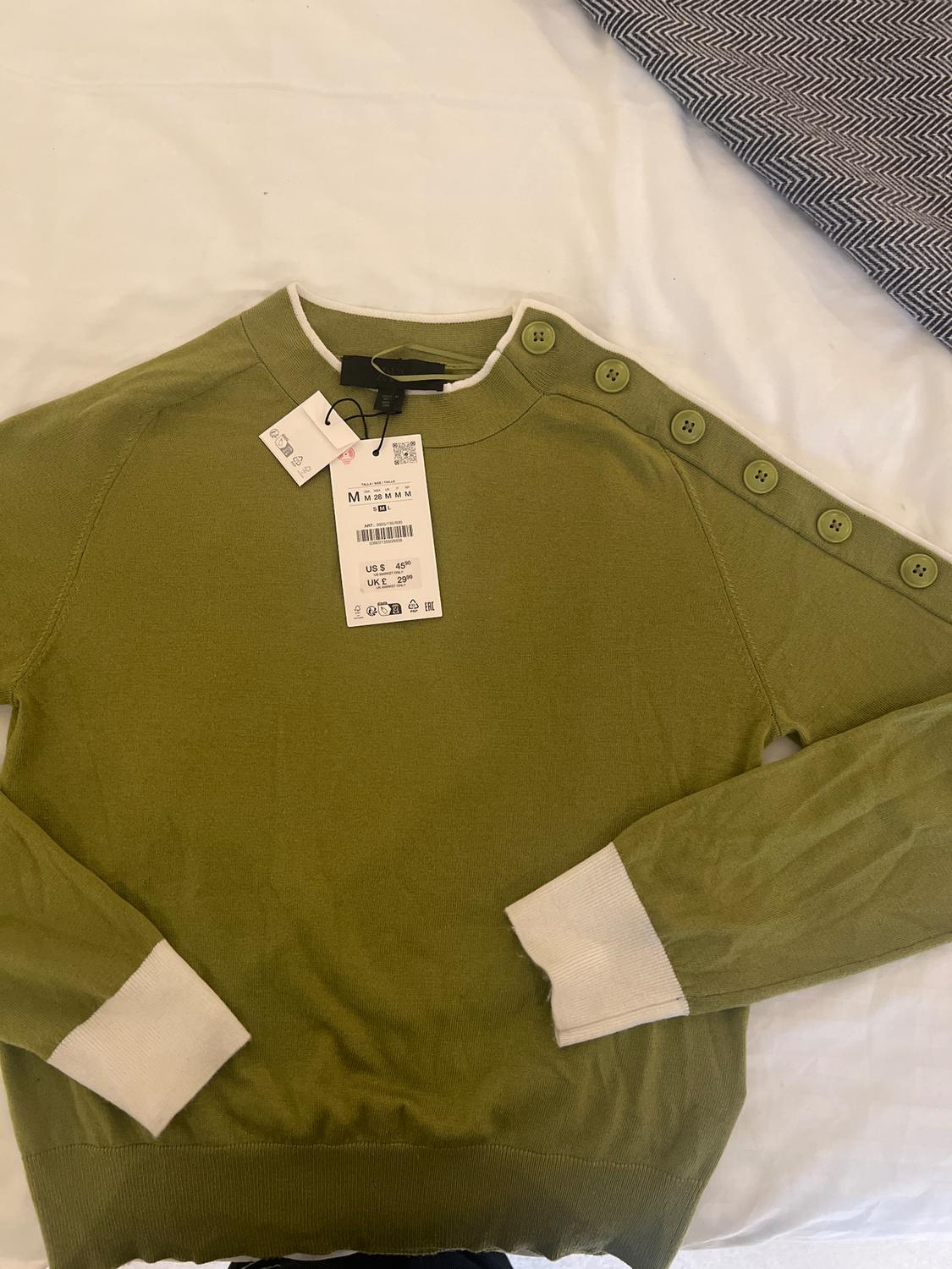 Olive green jumper with shoulder buttons and white cuffs. Visible tag with price of US$ 45.99 and size M. White fabric background.