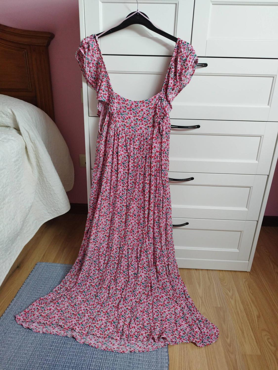 Pink long floral print dress hanging on a white piece of furniture in a bedroom with a bed, pink wall, and wooden floor.