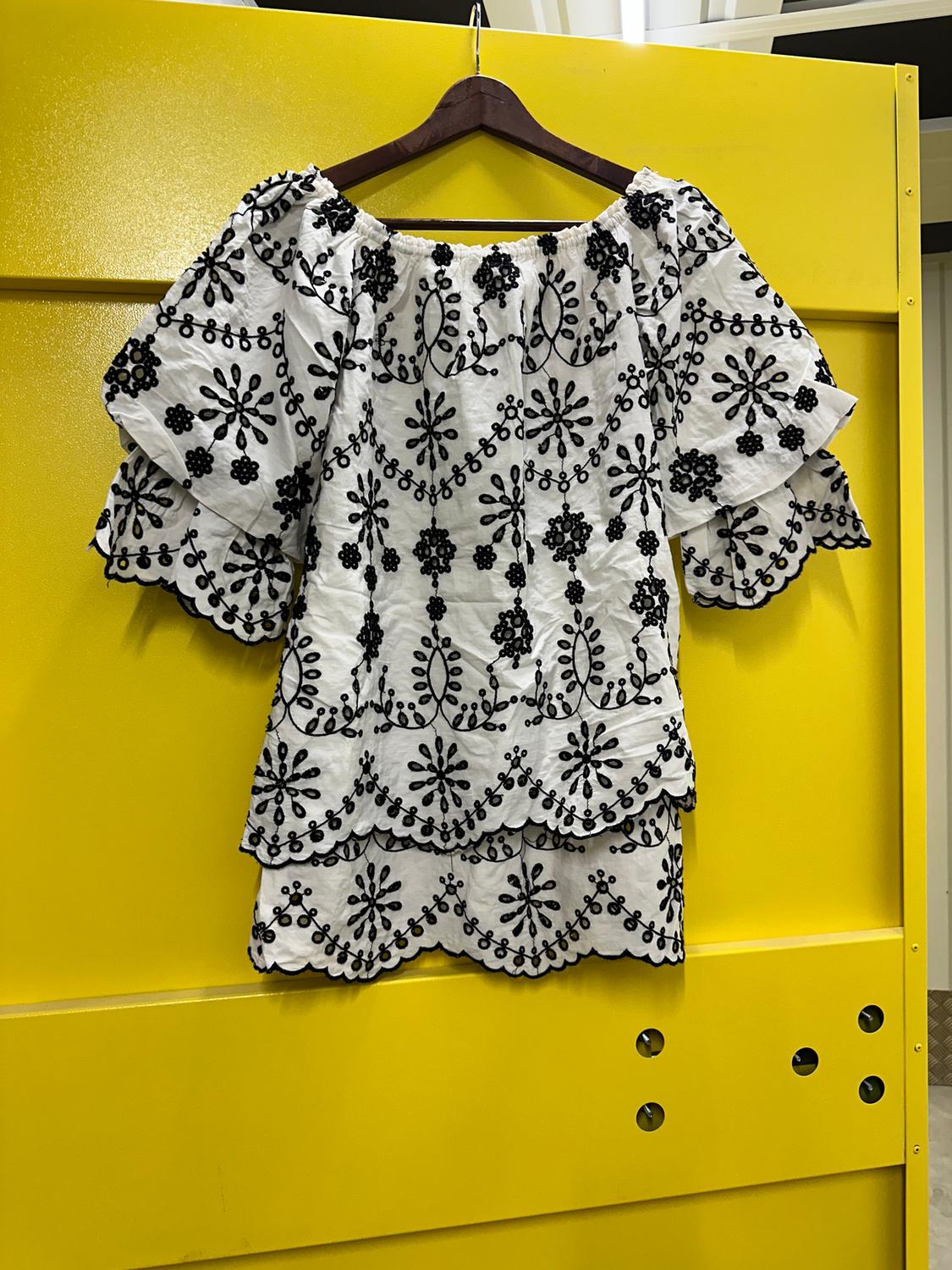 White blouse with black floral embroidery hanging on a wooden hanger against a bright yellow background.