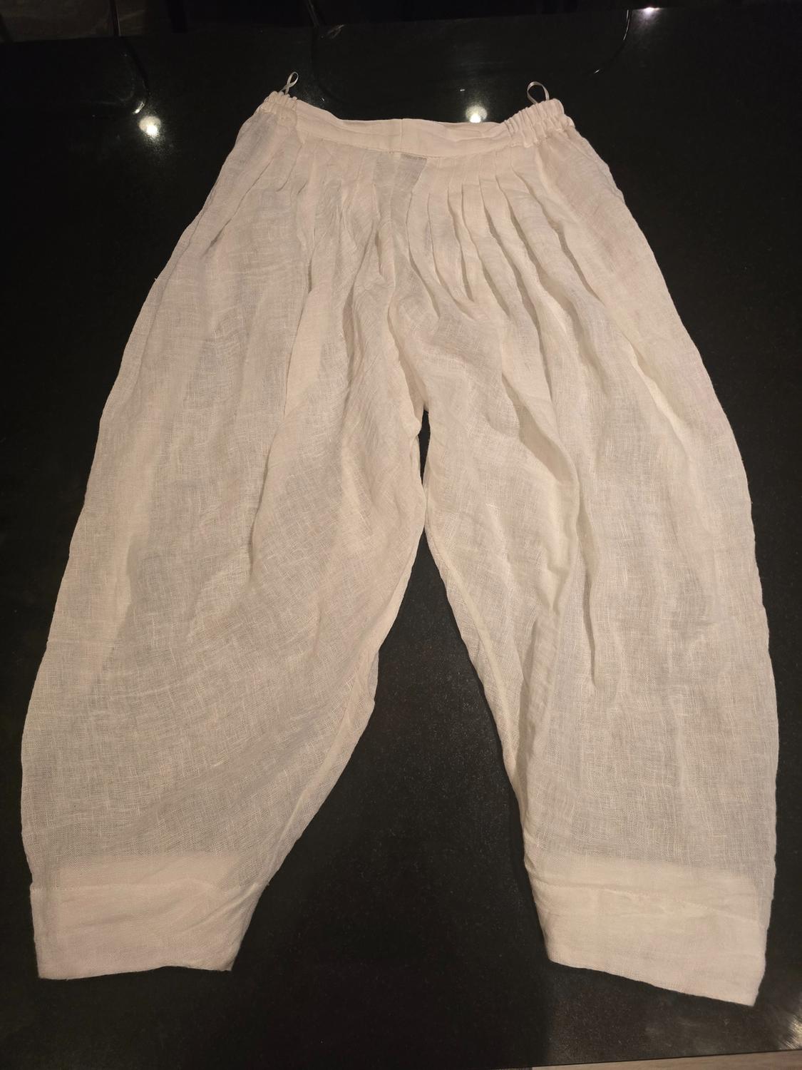 White linen pants with pleats, elastic waist, and wide cut, placed on a dark surface.