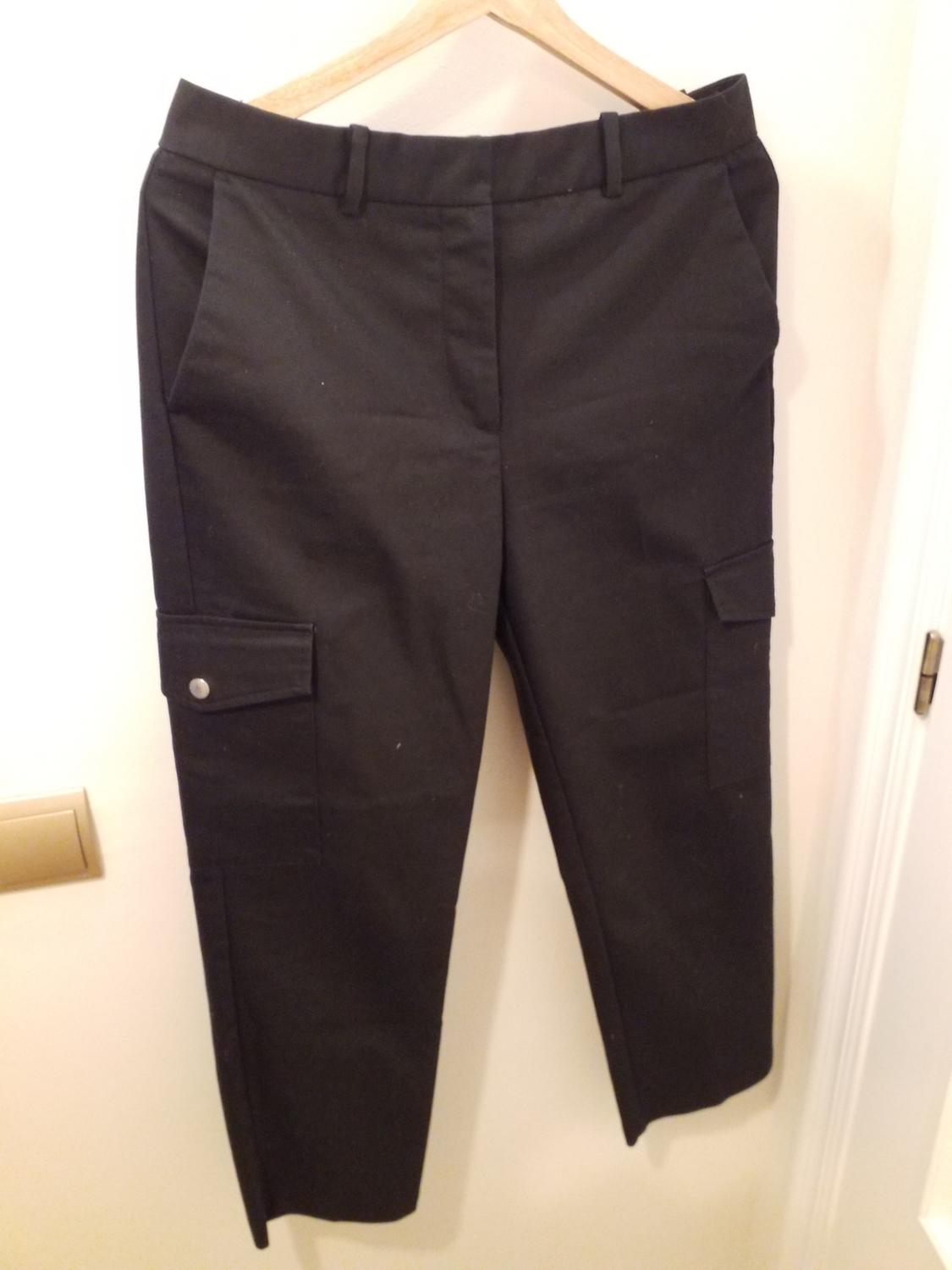 Black trousers hanging on a wooden hanger, with side pockets and button detail, on a white background.