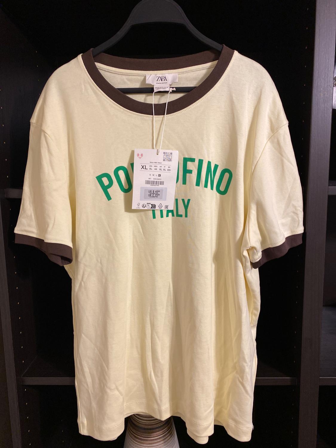 Beige t-shirt with brown collar and sleeves, "PORTOFINO ITALY" text in green. Visible hang tag. Hanging on a black clothes rail.
